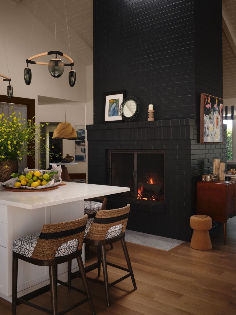 A modern kitchen with a black brick fireplace in the center, a fire burning inside. Three woven barstools with patterned cushions face a white island topped with a bowl of lemons and greens. Art, a clock, and flowers decorate the surrounding walls and surfaces.