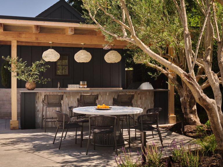 A stylish outdoor patio features a round white table with black wicker chairs, a bowl of oranges, and a shaded bar area with hanging woven lights. Mature trees, plants, and modern decor create a relaxed, inviting atmosphere in natural sunlight.