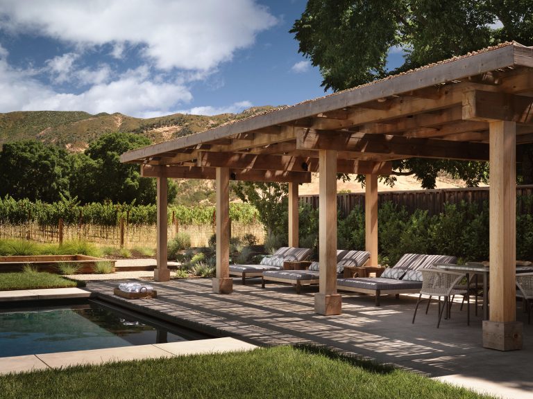 A wooden pergola shades cushioned lounge chairs and tables on a patio beside a pool. Lush green trees, a vineyard, and rolling hills are visible in the background under a blue sky with clouds. Sunlight casts shadows across the deck.