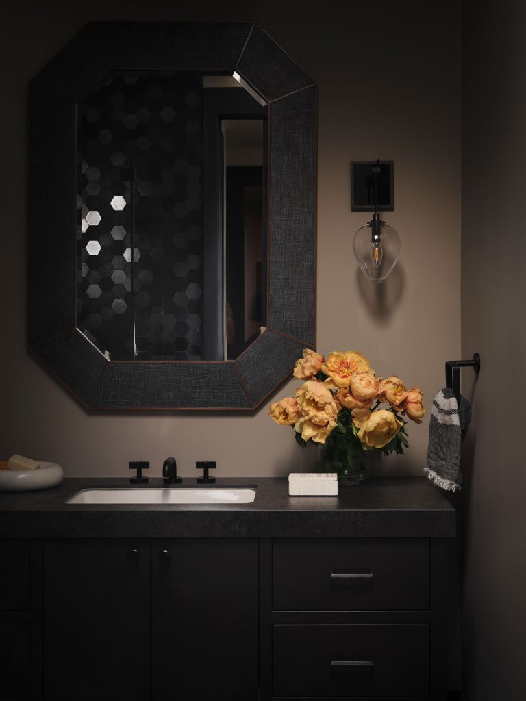 A modern bathroom with dark cabinetry and countertop features a black-framed octagonal mirror above a white sink. A vase of yellow-orange flowers, a hand towel on a ring, a wall light, and a bar of soap in a dish complete the cozy, elegant decor.