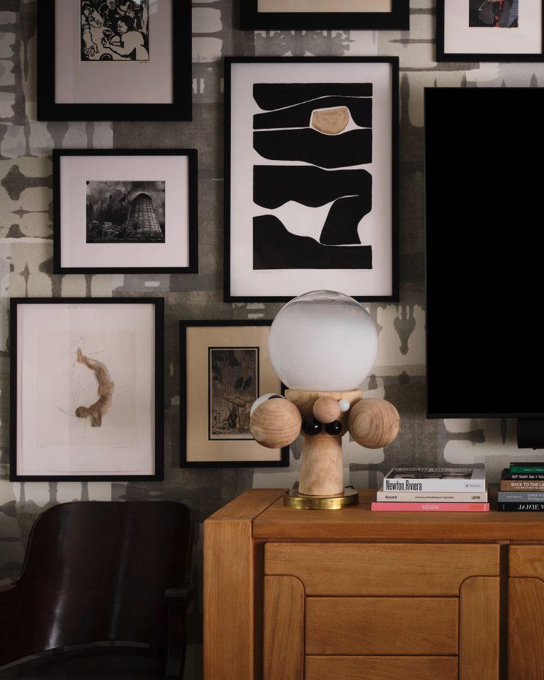 A modern interior features a wooden dresser with books and a unique lamp. Behind it, a collage of framed black-and-white abstract and photographic art hangs on a patterned gray wall. A dark wood chair sits nearby. The scene feels stylish and cozy.