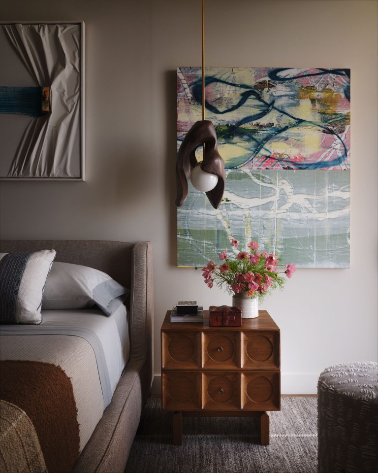 A cozy bedroom features a modern wooden nightstand with circular patterns, a vase of pink flowers, books, and a sculpture lamp. Above, abstract art hangs on the wall. Part of a bed with neutral bedding and a textured pouf are also visible.