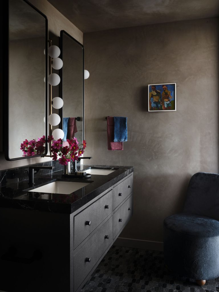 Modern bathroom with dark cabinetry and black marble countertop, featuring dual sinks and two large mirrors. Spherical vanity lights are mounted between the mirrors. Pink flowers in a vase, colorful towels, abstract wall art, and a dark plush chair add decorative touches.