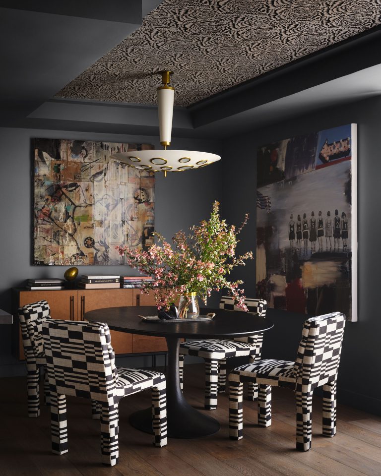 A modern dining room with black-and-white checkered chairs around a black table holding a vase of pink flowers. Abstract art hangs on dark walls; one painting is colorful, another has grayscale tones. A geometric ceiling pattern and a gold-accented pendant light are overhead.