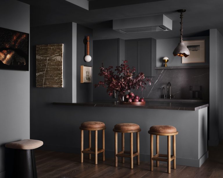 A modern kitchen with dark gray walls and cabinets, wooden bar stools with brown cushions, a marble-patterned backsplash, pendant lights, and autumnal red foliage arranged in a vase on the counter, creating a cozy, contemporary atmosphere.