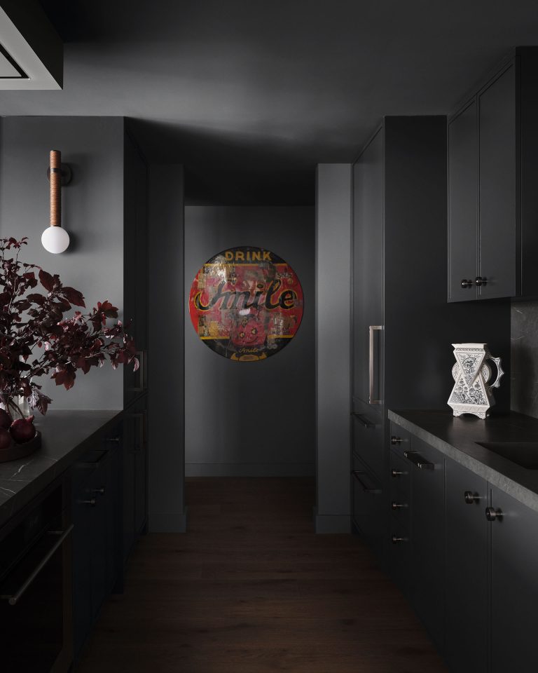 A dimly lit modern kitchen with dark gray cabinets and countertops. On the left, a vase with dark red leaves sits under a round wall sconce. In the hallway, a vintage red and yellow Drink Smile sign hangs on the wall, adding color to the space.