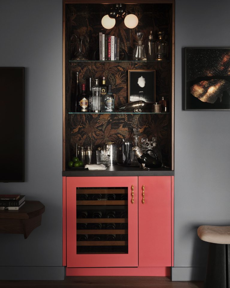A stylish home bar nook with coral pink cabinetry, gold handles, a glass wine fridge, and dark wallpaper with a leafy pattern. Shelves display glassware, bottles, books, and bar tools. A dark wall has framed art, and nearby furniture is partially visible.