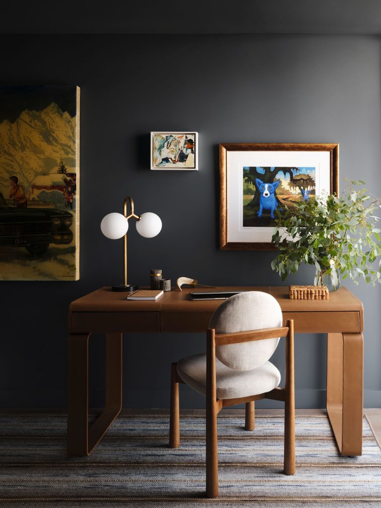A modern home office with a sleek wooden desk, round-cushioned chair, and gold lamp. Art hangs on a dark wall, including a painting of a blue dog. A leafy plant sits on the desk, which is tidy, with books and a notepad. A striped rug covers the floor.