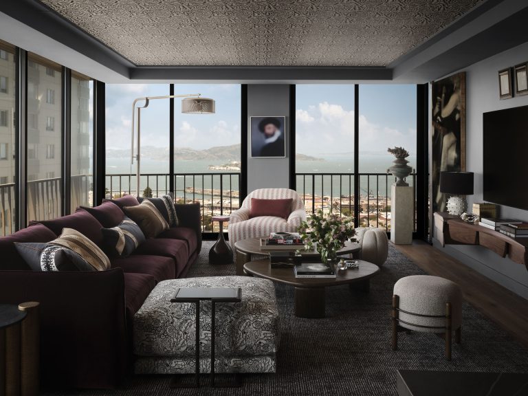 A modern living room with large windows overlooks a bay. The space features a burgundy sofa, patterned armchair, circular coffee tables, and dark decor. Art and plants decorate the room, and city buildings are visible outside under a bright blue sky.