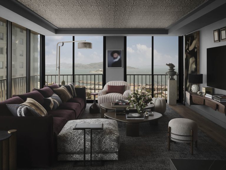 A stylish, modern living room with dark walls and a textured ceiling. Large windows reveal a scenic view of water and distant hills. The room is furnished with a maroon sofa, patterned armchairs, round coffee tables, and decorative plants, creating a cozy, elegant atmosphere.