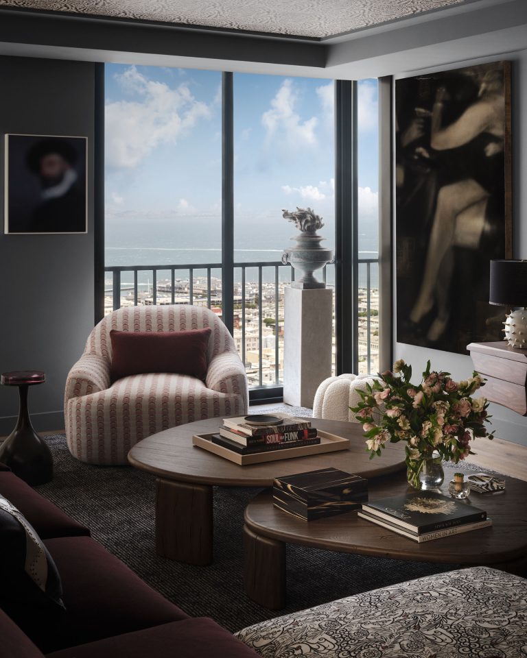 Modern living room with large windows and city and bay views. Room has patterned armchair, dark sofa, round wooden tables with books and a vase of flowers. Wall art and a statue on a pedestal are visible; room has neutral tones and soft natural light.