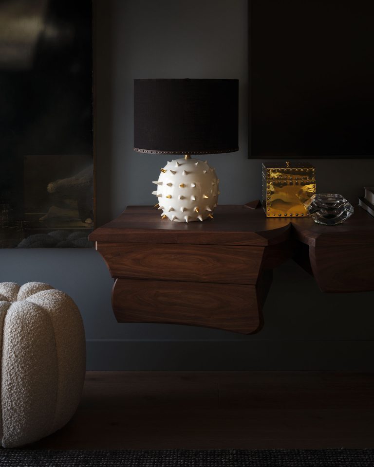 A modern interior with a floating wooden shelf holds a unique white lamp with gold spikes, a shiny gold box with studded accents, and a glass ornament. A textured, cream ottoman sits nearby and part of a dark painting and black wall are visible.