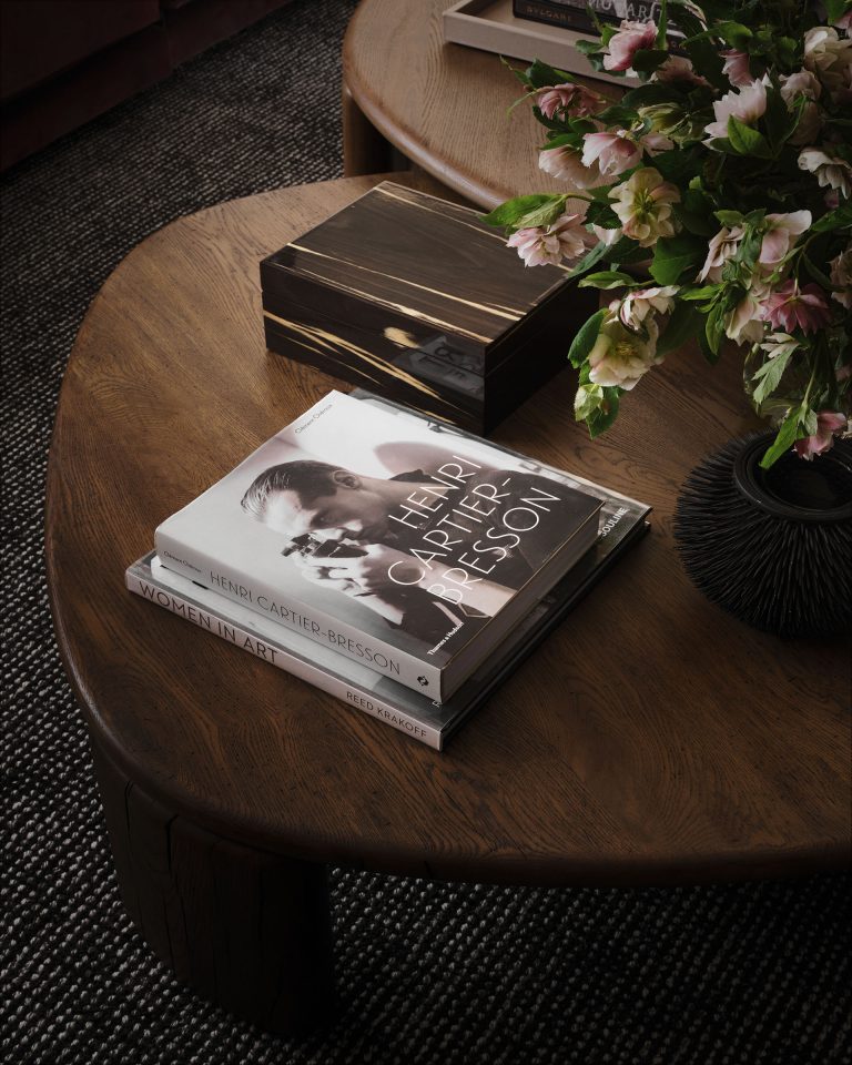A wooden coffee table holds stacked books, including Henri Cartier-Bresson with a black-and-white cover photo, Women in Art, and a book with a dark wood cover. A vase with pink and white flowers sits on the table next to the books.