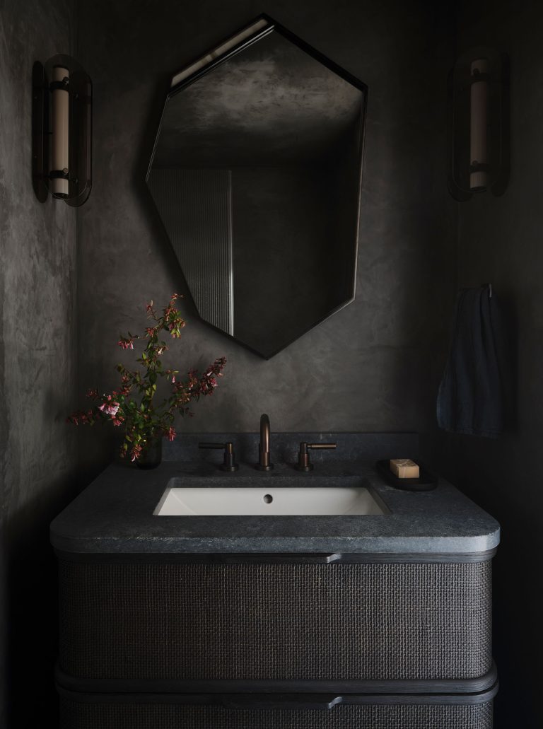 A modern bathroom with dark walls, features a rectangular sink set in a dark vanity. There is a black geometric mirror above the sink, two wall sconces on either side, a towel hanging on the right, and a vase with red flowers on the left side of the counter.