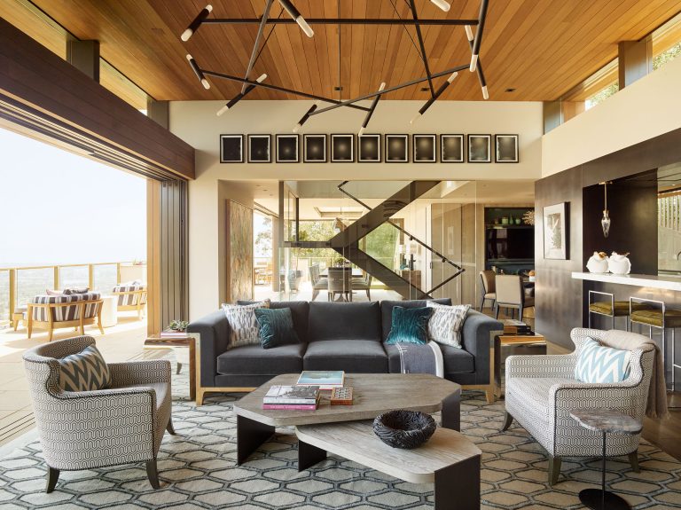 Modern living room with large windows and wood ceiling. Features a dark gray sofa, two patterned armchairs, geometric coffee tables, and a rug with hexagonal patterns. Abstract chandelier hangs overhead; glass doors open to a deck with a water view.