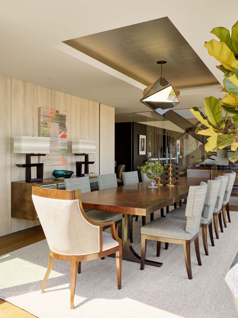 A modern dining room with a long wooden table, ten upholstered chairs, and a beige armchair at the head. Geometric pendant light hangs above. Art and decor on a sideboard, large leafy plant, and floor-to-ceiling windows brighten the space.
