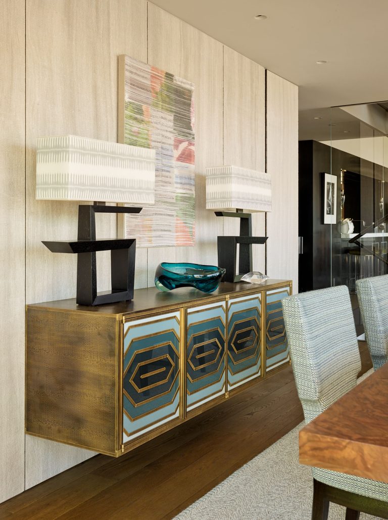 A modern dining room features a wooden sideboard with geometric blue and gold cabinet doors, topped with two black sculptural lamps with white patterned shades and a blue glass bowl. Textured beige walls and a colorful abstract art piece hang above.