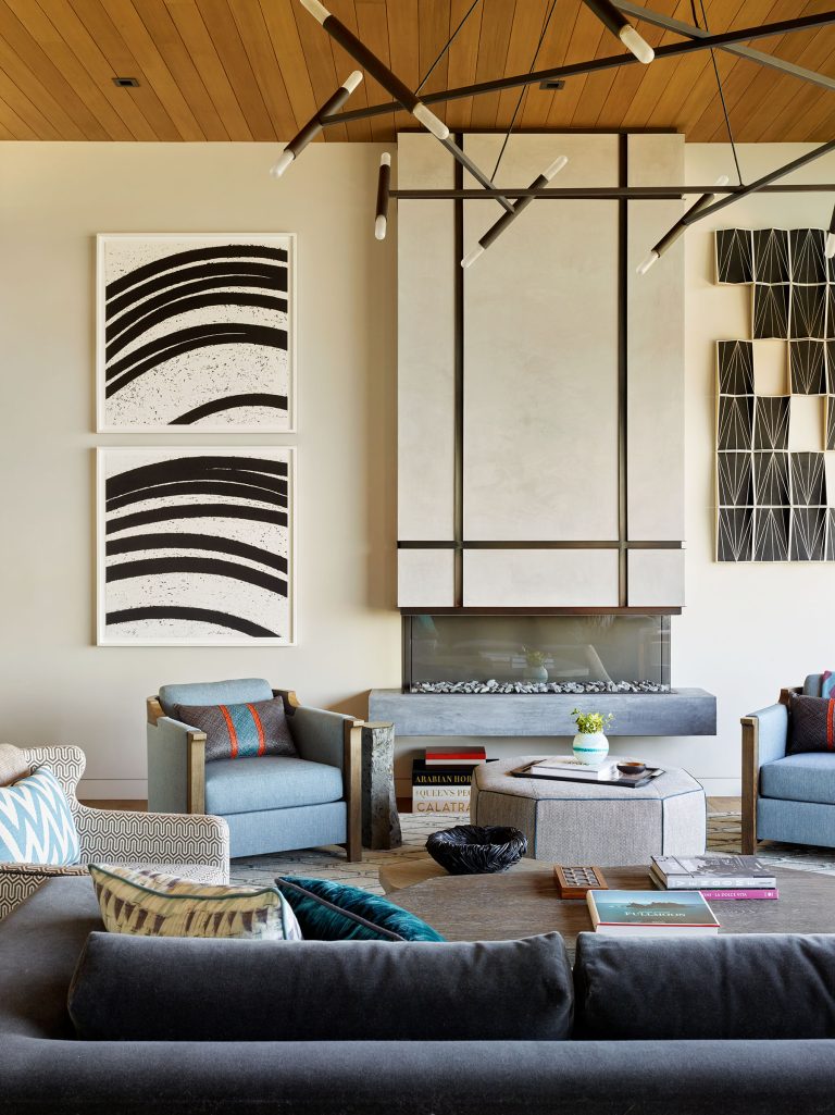 A modern living room with geometric decor features a sleek fireplace, abstract black-and-white art, patterned armchairs, a dark sofa, and a round ottoman with books. A striking black chandelier and wood-paneled ceiling add warmth and contemporary style.