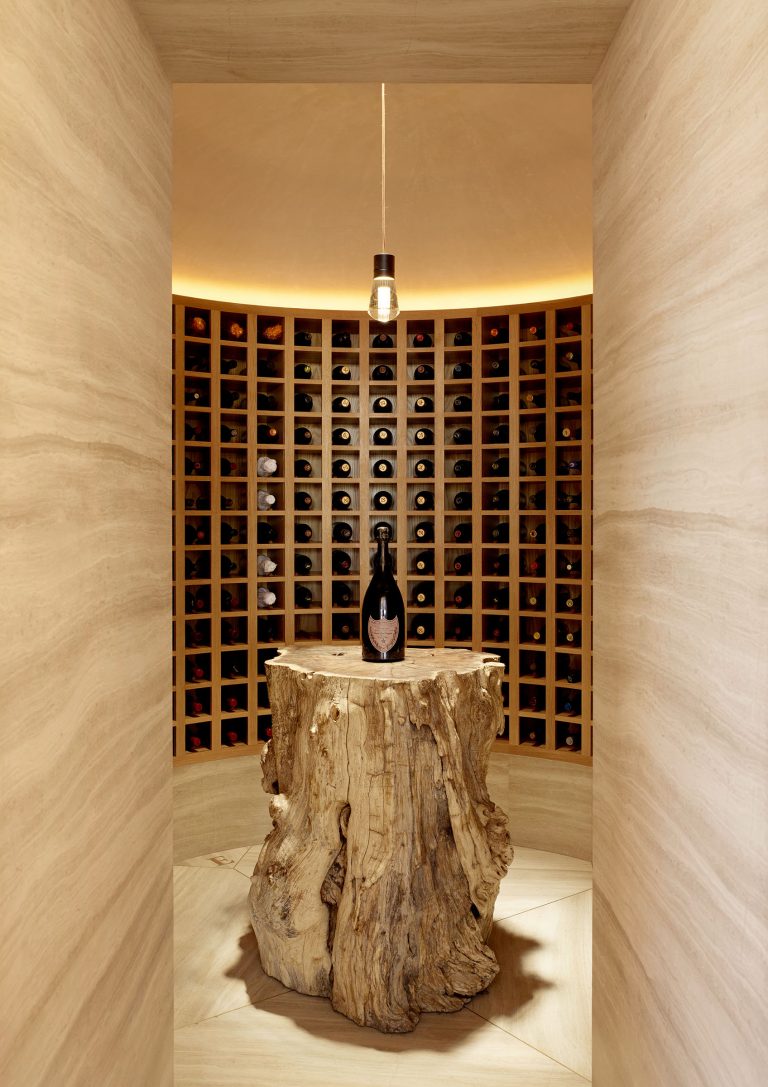 A single wine bottle sits atop a large, rustic tree stump pedestal in the center of a round, softly lit wine cellar, surrounded by curved shelves filled with neatly arranged wine bottles. A pendant light hangs overhead.