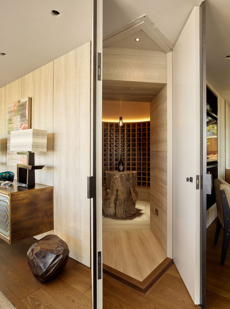 A modern wine cellar is hidden behind a white angular secret door in a wood-paneled wall. Inside, wine bottles are stored in a grid, and a sculpted tree stump serves as a table. Warm lighting highlights the sleek, elegant design of the cozy room.