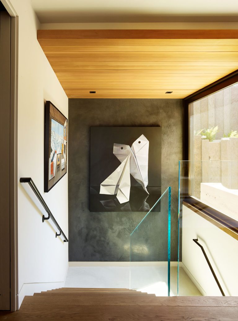 A modern staircase with wooden steps, a glass railing, and black handrails descends toward a dark accent wall featuring a geometric dog painting. Natural light streams in from a large window, illuminating the clean, contemporary space.