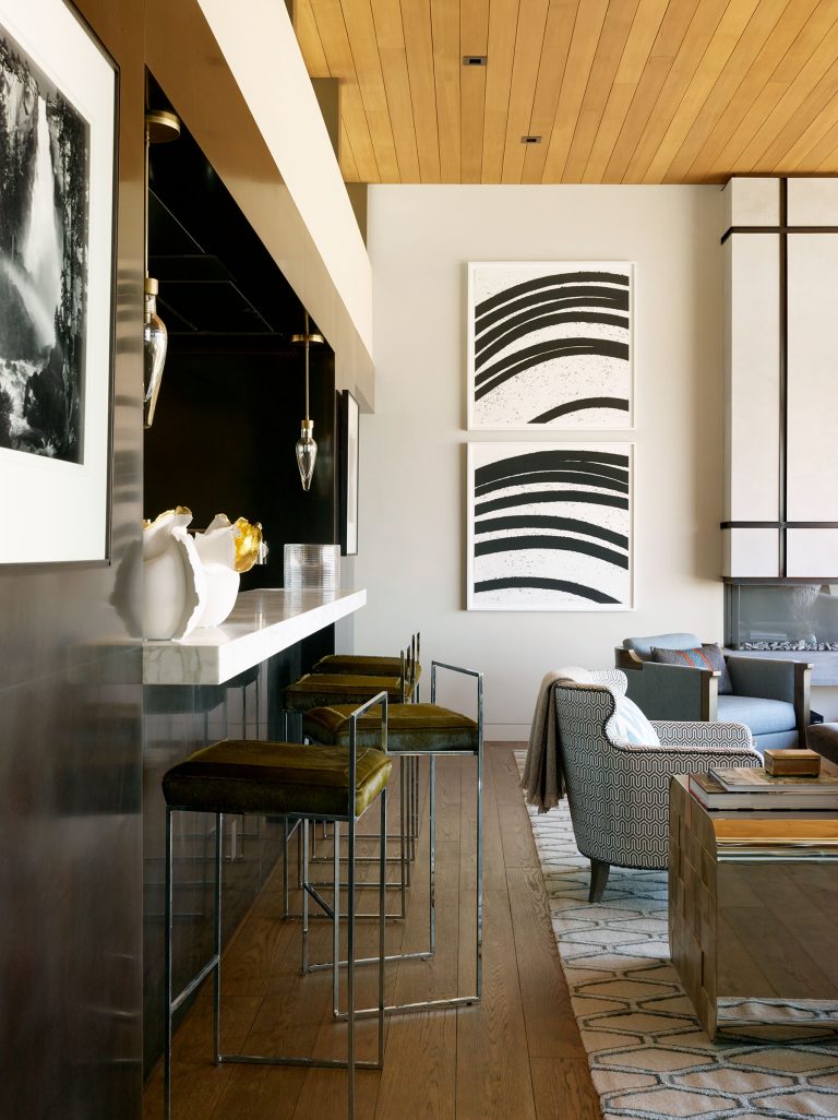 Modern open-plan living space with a wood ceiling and floor. A bar area with three velvet barstools sits beside abstract black-and-white wall art. The lounge area features patterned armchairs, a rug, and a fireplace. Monochrome décor creates a sleek, stylish look.