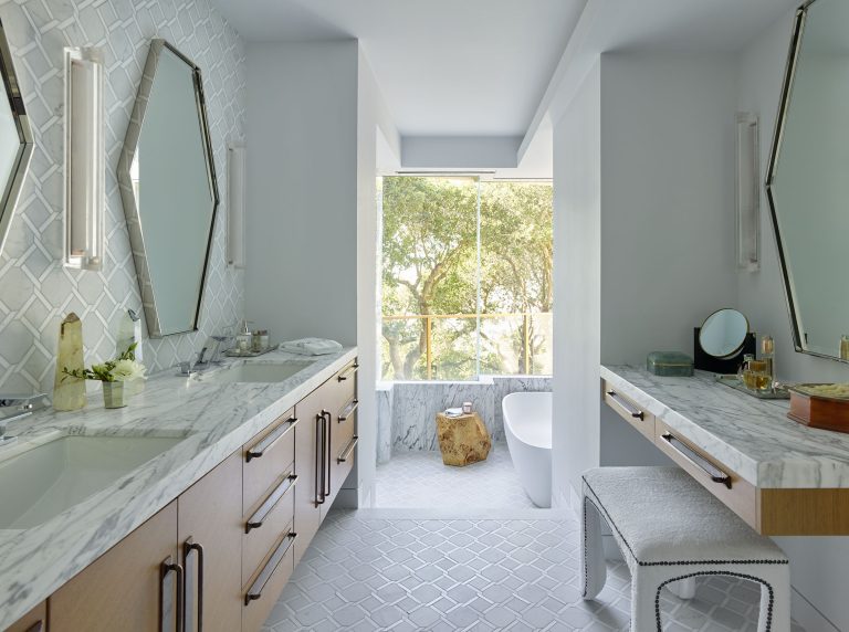 A modern bathroom with double sinks, marble countertops, light wood cabinets, two geometric mirrors, and wall sconces. A white freestanding tub sits by a large window with a tree view. The floor has a white tile pattern, and a vanity with a cushioned seat is on the right.
