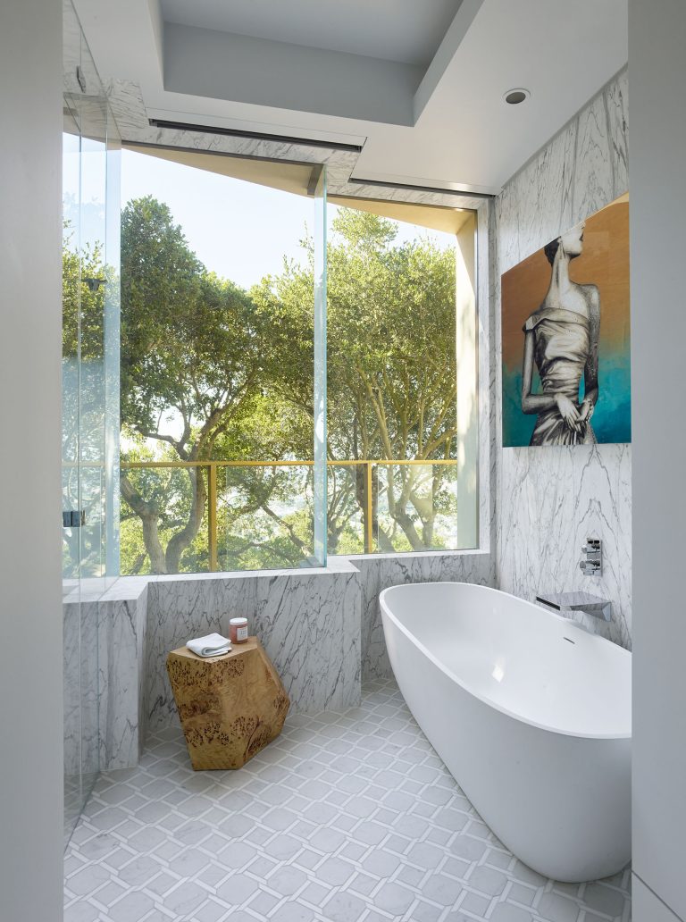 A modern bathroom with marble walls and floor, a freestanding white tub, a wooden side table holding a pink cup, and large windows revealing green trees outside. Abstract artwork in warm colors hangs on the wall above the tub.