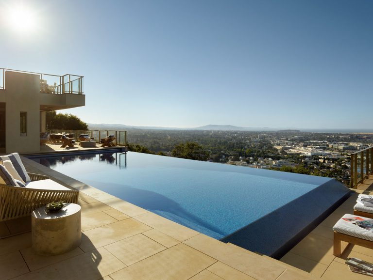 A modern infinity pool overlooks a sprawling cityscape on a sunny day. Lounge chairs sit by the pool, and a nearby building with glass balconies is visible. The sky is clear, and the city stretches into the distance under the bright sun.