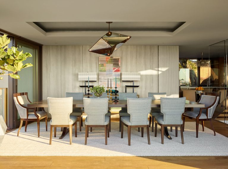 A modern dining room with a large wooden table surrounded by ten upholstered chairs on a light rug. There’s a geometric pendant light above, a plant to the left, built-in shelves, decorative objects, and a colorful artwork on the wall in the background.