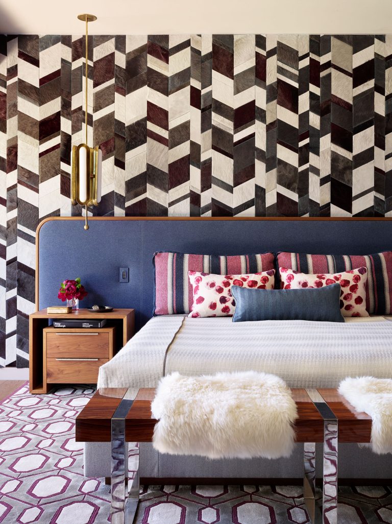 A stylish bedroom with a bold chevron-patterned accent wall in shades of brown, white, and gray. The bed has a blue upholstered headboard, striped and floral pillows, and a white bedspread. Wood benches with white fur throws sit at the foot of the bed.