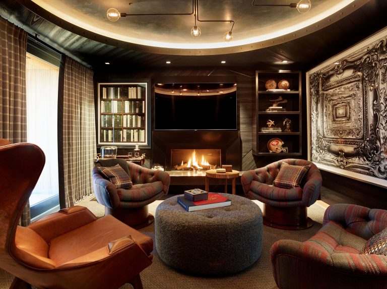 Cozy, modern lounge with four plaid swivel chairs around a round gray ottoman, a leather armchair, a lit fireplace below a large TV, bookshelves, sports memorabilia, and intricate monochrome wall art. Warm lighting and plaid curtains create an inviting atmosphere.