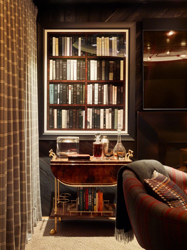 A cozy, dimly lit room features a framed photo of bookshelves, a mid-century bar cart with glassware and decanters, books underneath, a dark fireplace, plaid armchair with a blanket and pillow, and plaid curtains to the left.