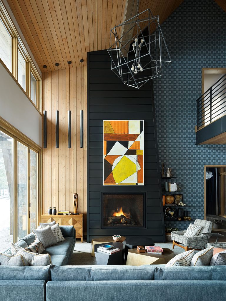 Modern living room with high wooden ceiling, large windows, and blue patterned accent wall. A black fireplace sits below abstract geometric art in yellow and orange. Blue sofas, a wooden coffee table, and a sculptural light fixture complete the cozy space.