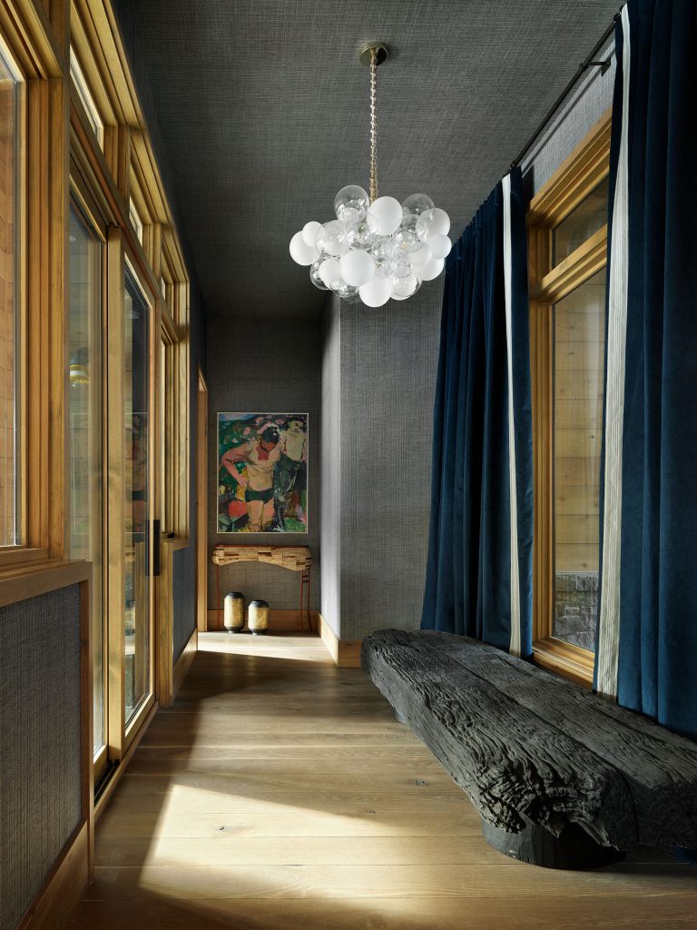 A narrow hallway features wood-framed glass windows, a dark textured wall, deep blue curtains, a rustic wooden bench, a modern bubble chandelier, a small console table, two decorative vases, and a colorful framed abstract painting on the far wall.