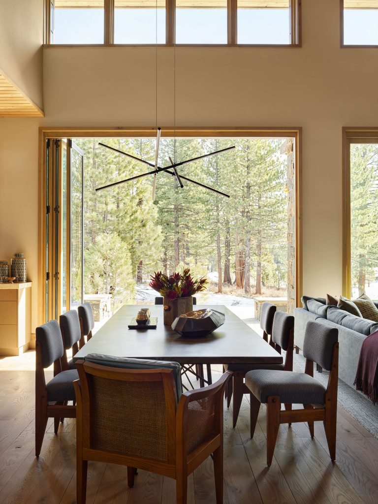 A modern dining room with a rectangular table, eight cushioned chairs, and a geometric black chandelier. Large glass doors open to a forest view, letting in natural light. A cozy living area with a gray sofa and cushions is partly visible to the right.