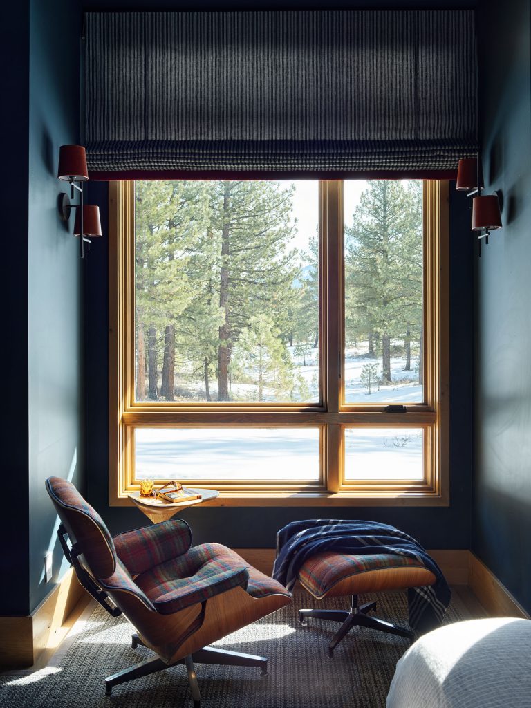 A cozy reading nook features a plaid armchair and footrest beside a window with snowy pine trees outside. A throw blanket drapes over the footrest, and a small table with a book and glasses sits nearby. The room has dark blue walls and soft natural light.