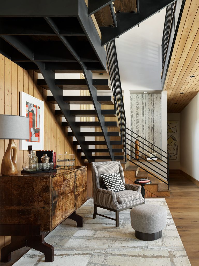 A modern living space features a black metal open-riser staircase with wood treads, light wood-paneled walls and ceiling, a patterned armchair with a round ottoman, a wooden sideboard with a lamp, art, and glassware, and a small desk beneath the stairs.