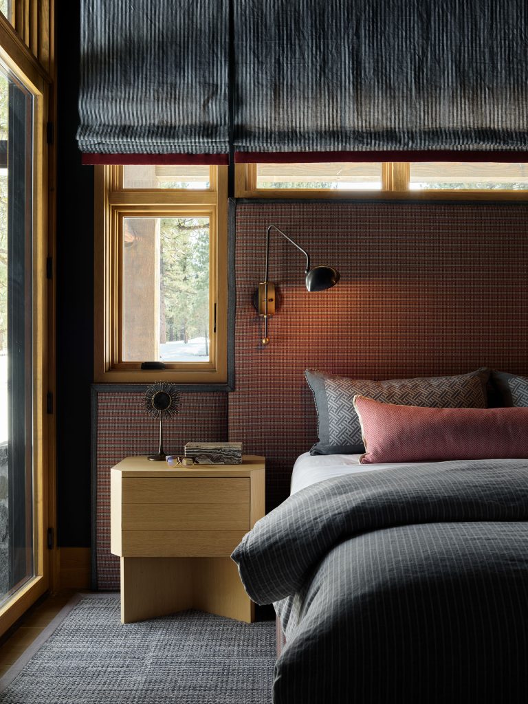 A cozy bedroom features a bed with gray bedding and mauve accent pillows. A wooden nightstand holds a small sculpture and lamp. Wall-mounted sconce lights the space. Large windows with textured blinds let in natural light; trees are visible outside.