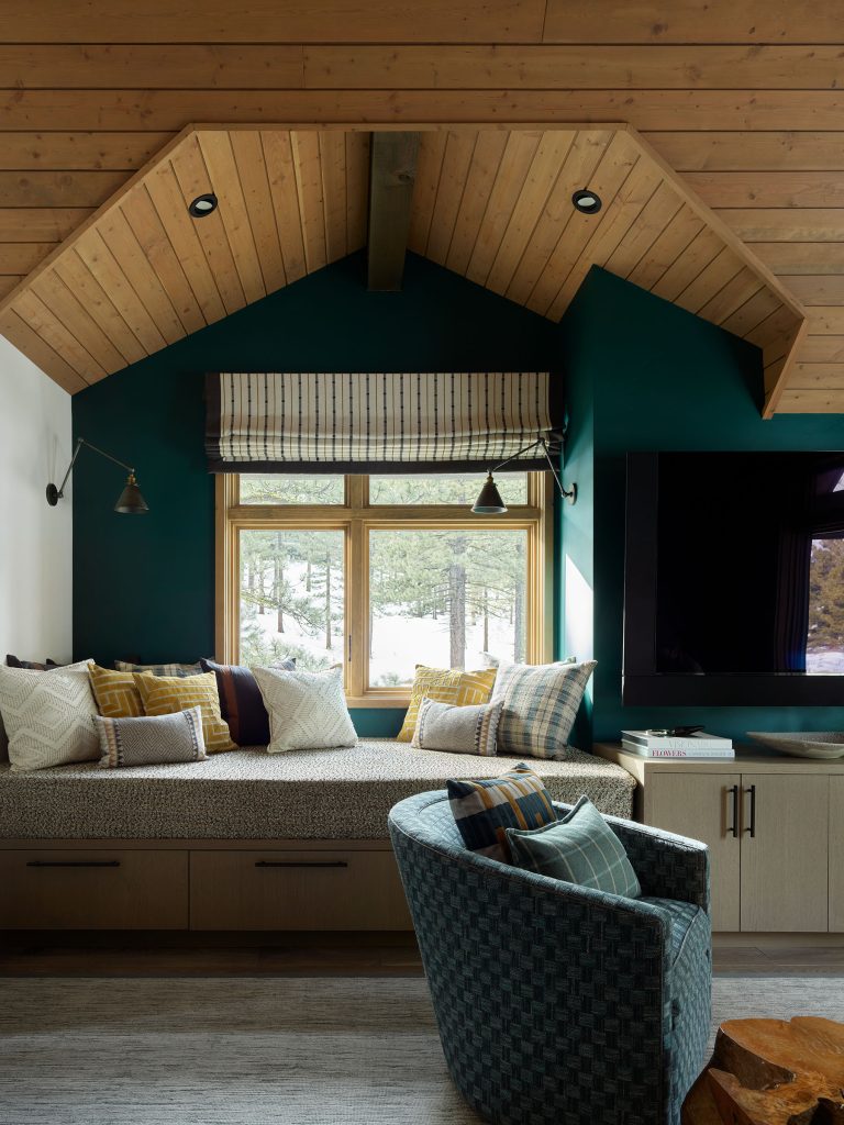 A cozy corner features a built-in window seat with gray cabinetry, white and patterned pillows, and a striped Roman shade. A plaid accent chair sits in front. The sloped wooden ceiling and dark green accent wall create a warm, inviting atmosphere. Large window shows trees.