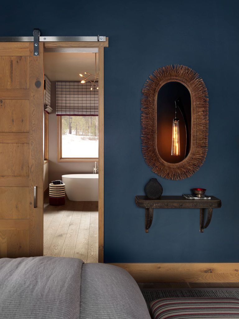 A cozy room with a blue wall, rustic wood sliding door, and a glimpse into a bathroom with a white bathtub. A woven-framed mirror with a hanging light, a small wall shelf holding decor, and a striped blanket on the bed are visible.