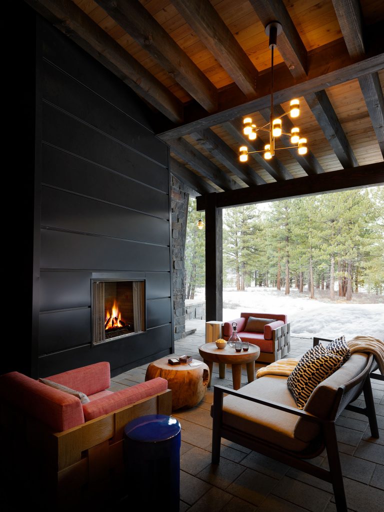 A cozy outdoor patio with modern wood furniture, red cushions, a round wooden table, and a roaring fireplace set in a sleek black wall. A contemporary chandelier hangs above. Large windows reveal snow and pine trees outside, evoking a warm, inviting winter atmosphere.