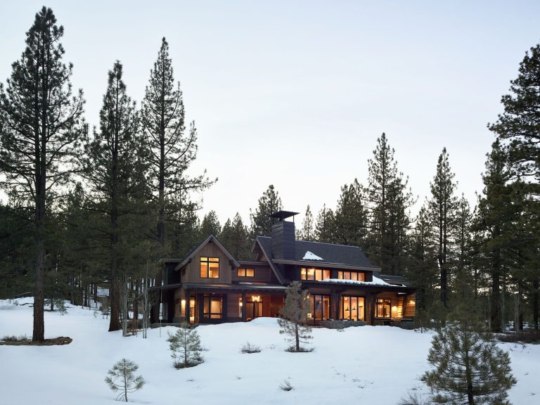 A modern dark-wood house with large windows glows warmly at dusk, surrounded by tall pine trees and a blanket of snow. The sky is pale and clear, creating a peaceful, wintry forest setting.