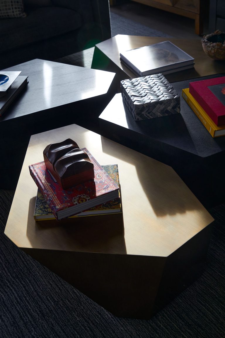A group of geometric, dark-colored coffee tables holds stacked books, a decorative box, and a dark abstract sculpture. Sunlight casts angular shadows across the tables, emphasizing their shapes and the textured surface beneath.