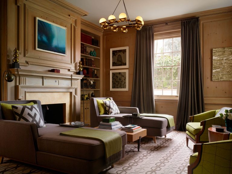 Elegant wood-paneled living room with two chaise lounges, green-accented pillows and throws, a fireplace, bookshelves, modern art on the walls, a large window with gray curtains, and a gold chandelier overhead. Natural light brightens the cozy, stylish space.