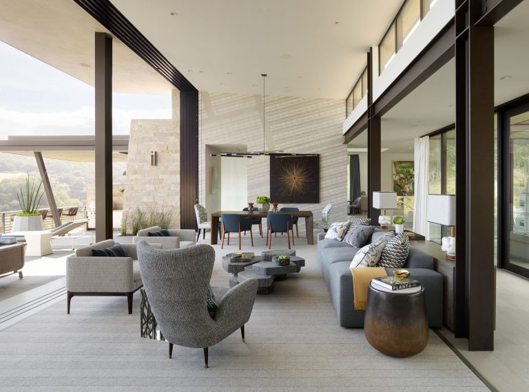A spacious modern living room with large windows, neutral-toned furniture, a gray sofa, armchairs, and a geometric coffee table. An open dining area is in the background, and outside views with hills and greenery are visible through the glass walls.