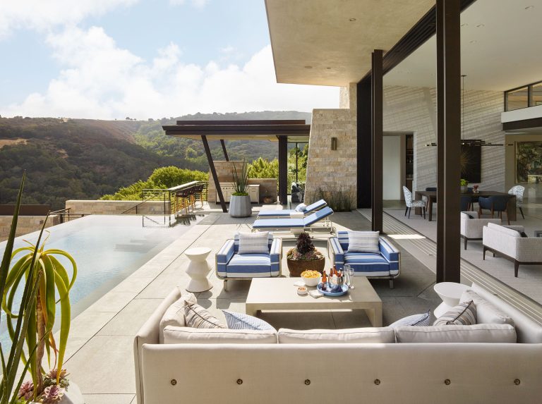 A modern outdoor patio with blue-striped lounge chairs, a beige sofa, and a pool overlooking green hills. There are drinks and snacks on the table, potted plants, and the house interior is visible through large sliding glass doors.