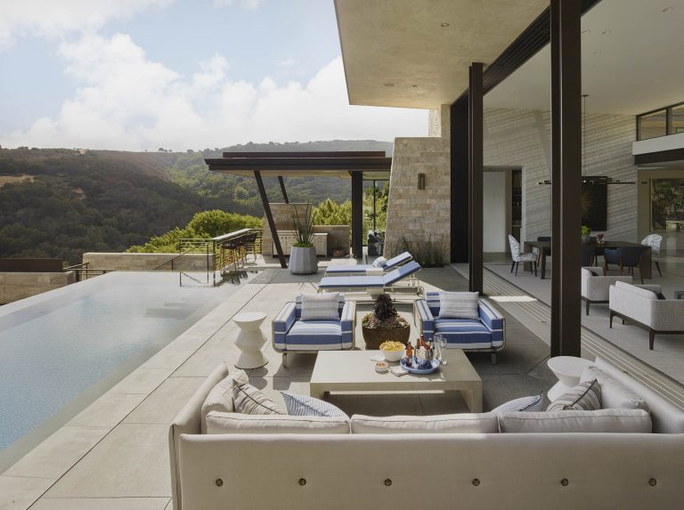 A modern outdoor patio with striped blue and white lounge chairs, a pool with an infinity edge, a beige sofa, a table with snacks and drinks, and scenic hills in the background under a partly cloudy sky. Large glass doors connect to an indoor living area.