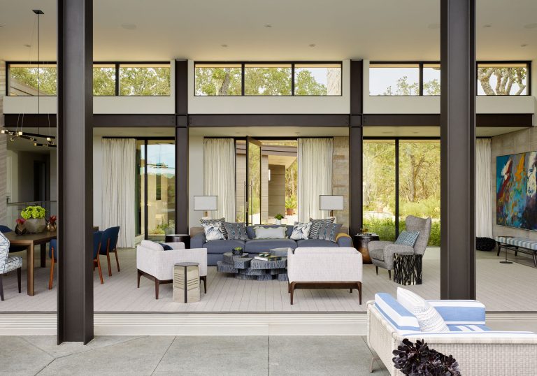A modern living area with floor-to-ceiling windows opens to a patio. The room features two sofas, multiple armchairs, a round coffee table, decorative pillows, and abstract art. Neutral tones, blue accents, and natural light create a bright, inviting atmosphere.