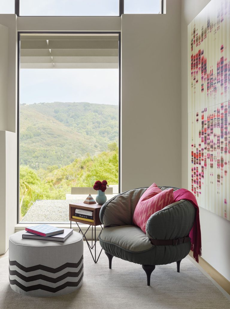 A cozy reading nook features a plush gray chair with pink pillows and a throw, a round ottoman with a chevron pattern, a small side table with books and a vase of dark pink flowers, large window with a view of green hills, and abstract art on the wall.